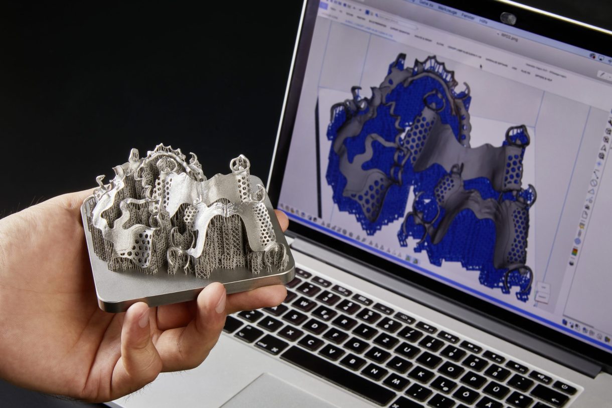 A hand holding a sample of 3d printed model in front of a 3d printed design on a laptop